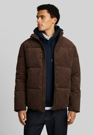 Man wearing a brown puffer jacket over a navy hoodie and white shirt, looking to the left against a plain gray background.