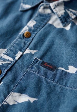 Blauwe denim overhemd met een wit vispatroon, een borstzak met een zwart label en bruine knopen op de sluiting. Gestructureerde stof.