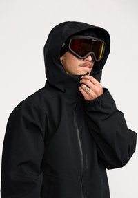 Veste noire avec capuche, dotée d'une fermeture éclair frontale et de poignets élastiques. Portée avec de grandes lunettes de ski, mettant en avant un design minimaliste.