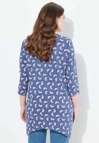 Blaue Tunika mit asymmetrischem Saum, ausgestattet mit einem weißen abstrakten Muster. Hergestellt aus weichem Stoff mit Dreiviertelärmeln.