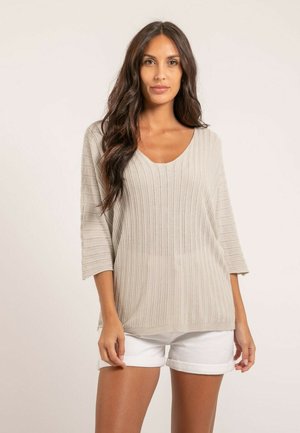Femme aux longs cheveux ondulés portant un pull beige côtelé à manches trois-quarts et un short blanc retroussé, debout devant un fond uni.