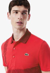 Röd polo med en gradientdesign, mörkröd krage och subtilt mönster. Inkluderar en grön krokodillogotyp på bröstet.