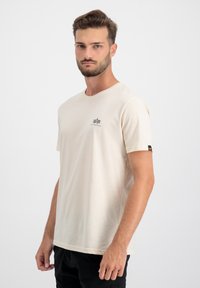 Beige Baumwoll-T-Shirt mit kurzen Ärmeln, rundem Ausschnitt und einem kleinen schwarzen Logo auf der Brust. Der Saum ist gerade, mit einem Etikett am Ärmel.
