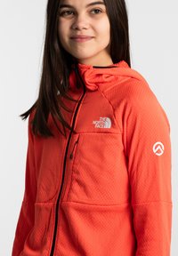 Giacca rossa con cappuccio realizzata in materiale trapuntato, con una tasca frontale e un logo sul petto e sulla manica. Zip e dettagli neri.