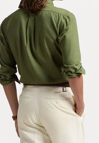 Zelená lněná košile s ohrnutými rukávy, zobrazena zezadu. Spárována s krémovými kalhotami s zadními kapsami a páskem.