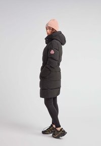Abrigo acolchado negro con cintura ceñida, capucha y parche de logo rosa; llevado sobre leggings negros y combinado con zapatillas negras y amarillas.