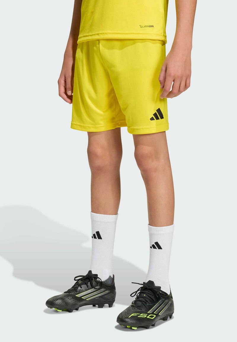 Enfant portant un short et un maillot de sport jaunes, des chaussettes blanches Adidas et des chaussures de football noires Adidas sur un fond uni.