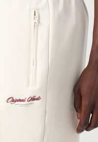 Shorts beiges en tissu doux avec une poche zippée sur le côté. Le nom de la marque est brodé en marron sur l'ourlet.