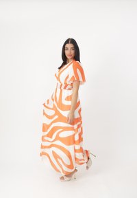 Maxikleid in Orange und Weiß, mit wellenförmigen Mustern, kurzen schwingenden Ärmeln und einer gerafften Taille. Das Model trägt hohe Sandalen.