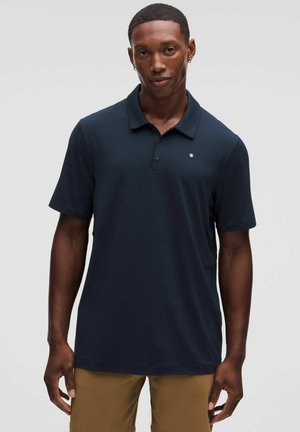 Giovane uomo con capelli corti che indossa una polo blu navy e pantaloni marroni, in piedi davanti a uno sfondo chiaro grigio uniforme.