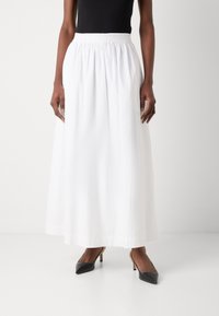 Jupe longue blanche avec un taille élastique, fabriquée à partir d'un tissu lisse. Associée à des talons noirs à bout pointu, soulignant un design épuré et minimaliste.