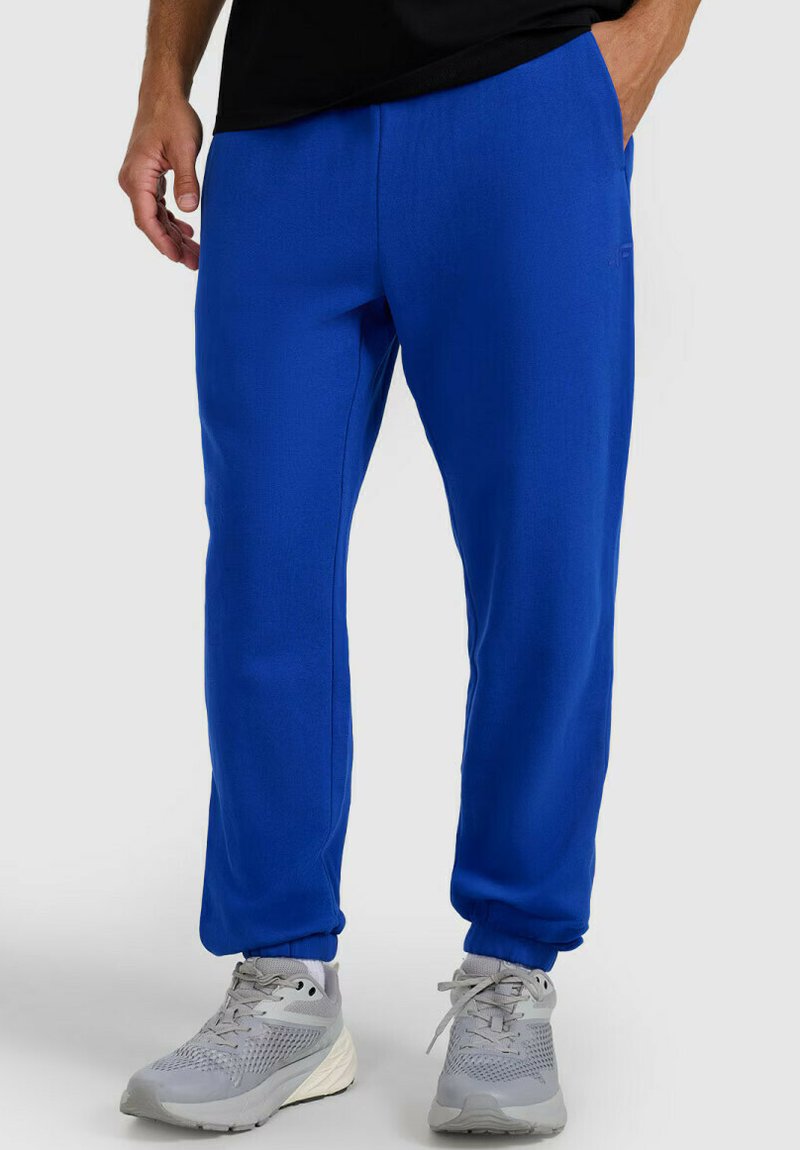 Mężczyzna w jasnoniebieskich spodniach dresowych z elastycznymi ściągaczami i szarych butach sportowych, stojący na jednolitym tle.