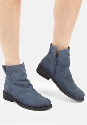 Füße, die mittelhohe blaue Wildleder-Stiefeletten mit seitlichen Reißverschlüssen und niedrigen schwarzen Absätzen auf weißem Hintergrund tragen.