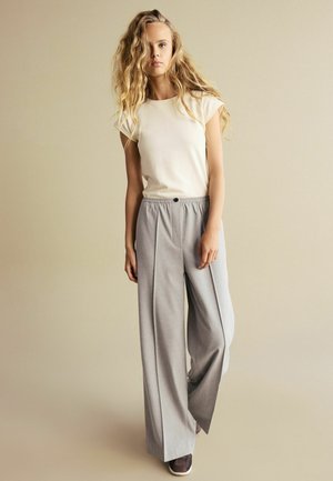 Beige tričko s krátkým rukávem v kombinaci s šedými kalhotami s vysokým pasem a širokými nohavicemi, které mají detail knoflíku a texturovanou látku.