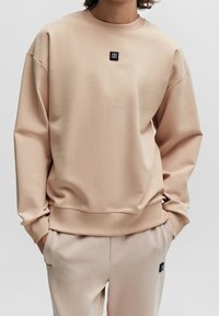 Sudadera de color beige claro hecha de una tela suave, con cuello redondo, hombros caídos y un pequeño parche negro del logo en el pecho.