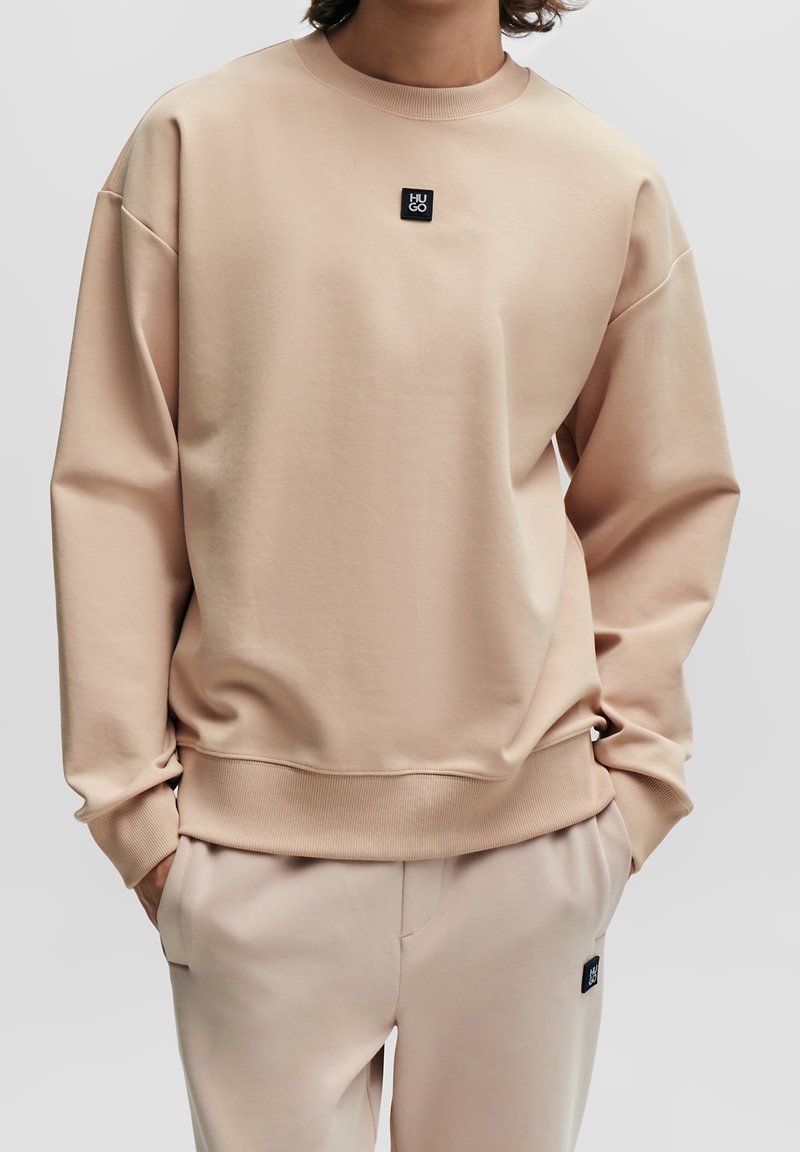 Sudadera de color beige claro hecha de una tela suave, con cuello redondo, hombros caídos y un pequeño parche negro del logo en el pecho.