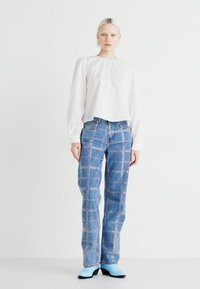 Blouse blanche à manches longues avec épaules froncées, associée à un jean en denim bleu clair avec un motif à carreaux et des embellissements métalliques.