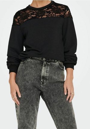 Person trägt ein schwarzes Langarmshirt mit floralen Spitzenakzenten auf den Schultern und hoch taillierte, ausgewaschene schwarze Jeans, die Hände ruhen an den Seiten.