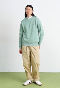 Felpa verde menta con polsini a coste, pantaloni beige a gamba larga e sneakers bianche con dettagli neri. Il modello si trova su un pavimento verde.