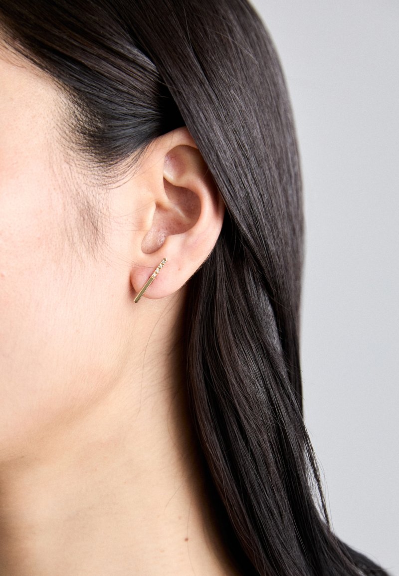 Χρυσά σκουλαρίκια με σχεδίαση στενής ράβδου, κοσμημένα με μια σειρά μικρών διακοσμητικών λίθων. Φοριούνται σε τρυπημένο αφτί.