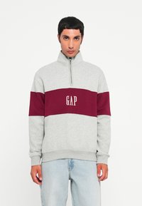 Gray sweatshirt with a maroon horizontal band, featuring a quarter zip and "GAP" logo in white. Soft fabric, relaxed fit.