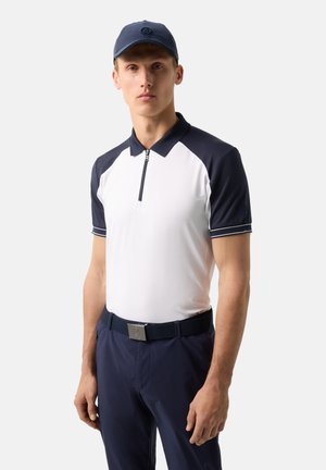 Mand iført navyblå kasket, hvid og navy zip-up poloshirt, navy bukser og bælte, stående mod en ensartet lys baggrund.