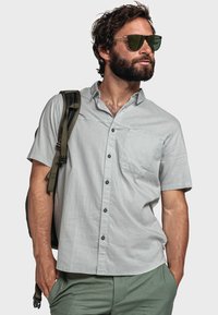 Kurzärmeliges, hellgraues Hemd mit Knopfverschluss und Brusttasche, schwarzen Knöpfen und einer subtilen Textur, getragen mit olivgrünen Hosen.
