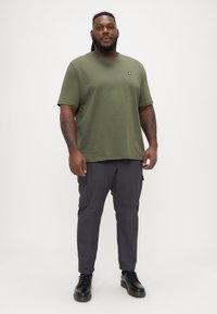 Tricou din bumbac verde măsliniu cu guler rotund și un mic logo galben. Asortat cu pantaloni cargo gri închis și pantofi negri cu șireturi.