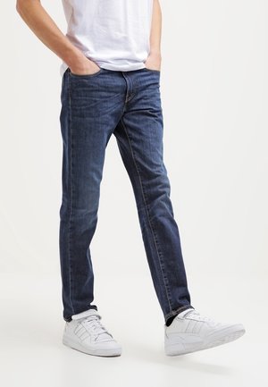 Jean bleu en denim avec une coupe slim, présentant un design classique à 5 poches et des coutures contrastantes. Associé à des chaussures de sport blanches.