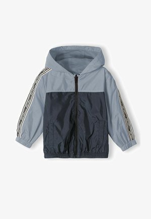 Lengvas, su gobtuvu vėjui atsparus paltas mėlynos ir tamsiai mėlynos spalvos. Jame yra užtrauktukas priekyje, elastingi rankogaliai ir šoninės kišenės su kontrastingomis šoninėmis juostelėmis.