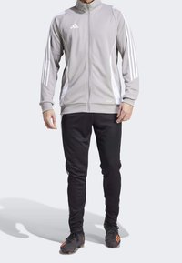 Graue sportliche Zip-Jacke mit weißen Seitenstreifen und einem Adidas-Logo, kombiniert mit schwarzen Hosen und schwarzen Stollenschuhen mit orangefarbenen Akzenten.