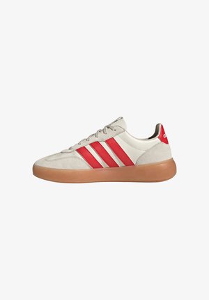 Off-white sneaker met gumzool, rode Adidas drie strepen aan de zijkant en een rood hielstuk met wit Adidas-logo.