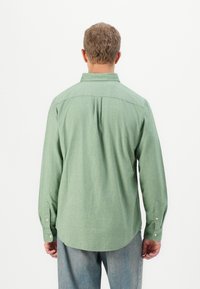 Hombre con cabello corto, vistiendo una camisa de manga larga verde claro y jeans azules, de pie frente a un fondo blanco.