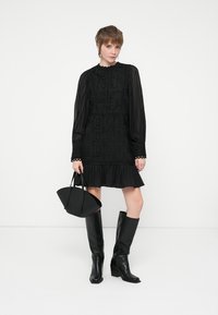 Musta pitsimekko pitkillä hihoilla, frillalla varustetun helman ja monimutkaisilla yksityiskohdilla. Yhdistetty mustiin polvipitkiin saappaisiin ja rakenteelliseen käsilaukkuun.