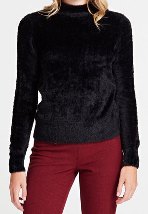 Femme portant un pull noir duveteux à manches longues et un pantalon rouge à carreaux, vue du cou aux hanches sur fond blanc.