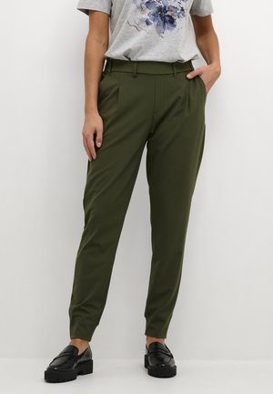 Persoon draagt olijfgroene taps toelopende broek, lichtgrijs bloemen T-shirt en zwarte chunky loafers, staat met één hand in de zak.