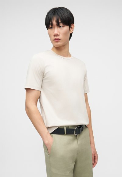 Νεαρός άνδρας με κοντά μαύρα μαλλιά, φοράει μπεζ κοντομάνικο t-shirt και ανοιχτοπράσινο παντελόνι με μαύρη ζώνη, τα χέρια στις τσέπες, κοιτάζοντας πλευρικά.