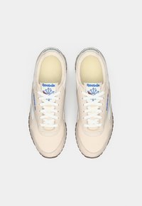 Bēšas sporta kurpes ar baltām auklām, mīksta auduma virspuse, mīkstināta apkakle un gumijas zole. Uz mēles ir zila Reebok zīmola identifikācija.