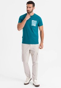 Teal cotton t-shirt with a light blue patterned pocket. Paired with light beige cargo pants and white sneakers with brown accents.
