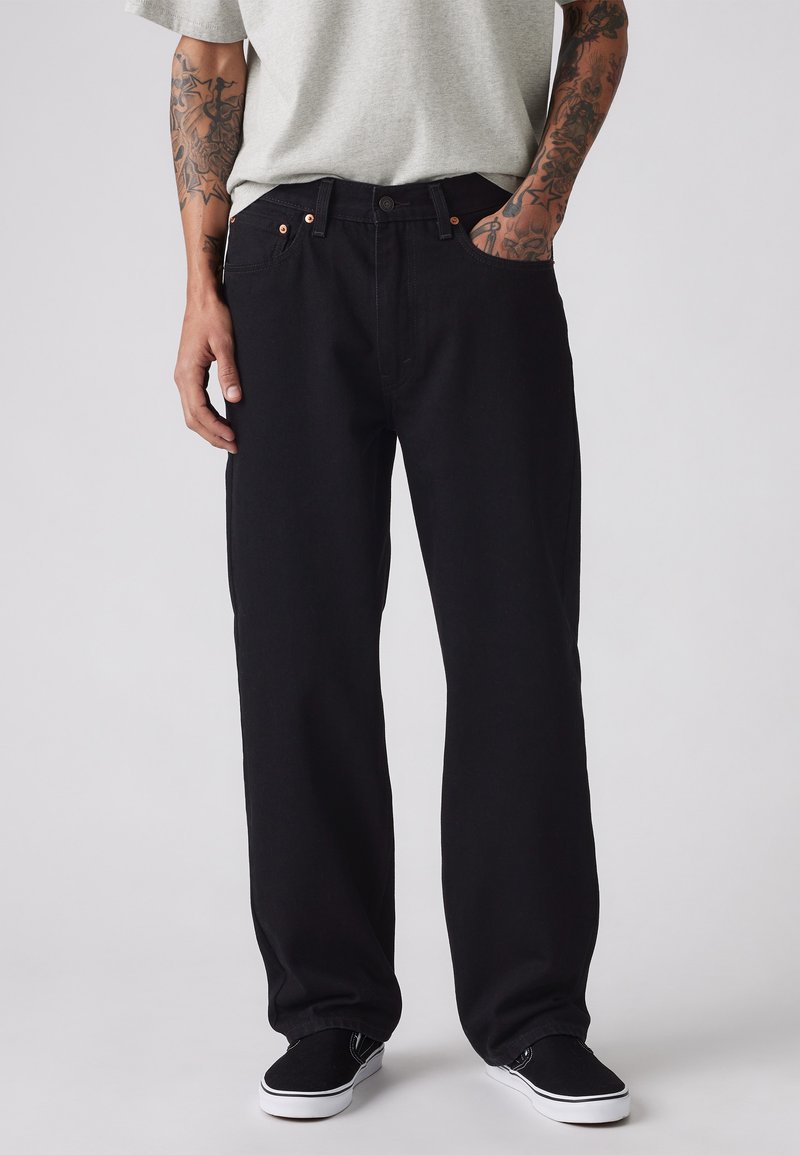 Pantaloni din denim negri cu o croială relaxată, având cuie de cupru pe buzunare, cusături decorative și asortați cu adidași negri.