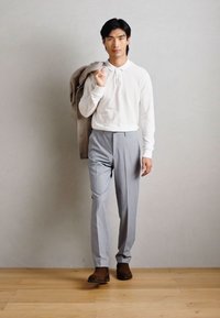 Άνδρας με λευκό μακρυμάνικο πόλο και γκρι παντελόνι περπατάει, κρατώντας μπεζ σακάκι στον ώμο, καφέ μπότες, ουδέτερο φόντο.