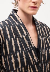 Jachetă din fabrică neagră cu un model abstract crem, are un guler lat și o decupare în formă de V, evidențiind detalii texturate ale suprafeței.