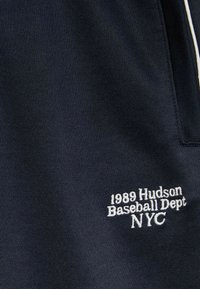 Granatowa tkanina sportowa o teksturowanym wykończeniu. Zawiera białe haftowane napisy: "1989 Hudson Baseball Dept NYC" w stylowej czcionce.