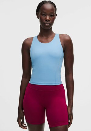 Kvinde iført en tætsiddende lyseblå ærmeløs top og højtaljede mørkerøde cykelshorts, der står mod en ensfarvet baggrund.