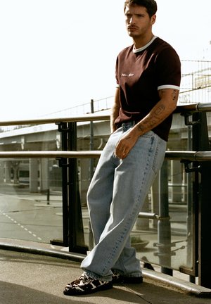 Mann mit Tattoos, der ein dunkles T-Shirt, hellblaue Jeans und gemusterte Schuhe trägt, lehnt draußen an einem städtischen Geländer.