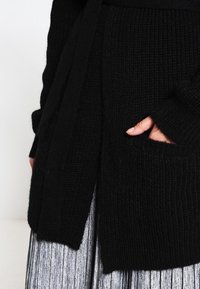 Ruka v kapse černého strukturovaného pleteného kardiganu nošeného přes metalickou plisovanou sukni, na světlejším jednobarevném pozadí.