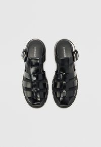 Sorte flette sandaler med glansfull overflate, som har justerbar spenne og teksturert såle. Åpen design og flat hæl.