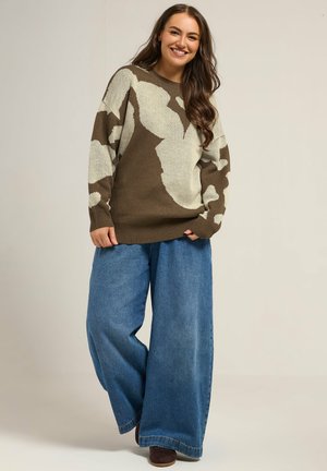 Übergroßer gestrickter Pullover in Braun mit cremefarbenem abstraktem Design, kombiniert mit weit geschnittenen blauen Denim-Jeans. Das Material wirkt weich und hat eine entspannte Passform.