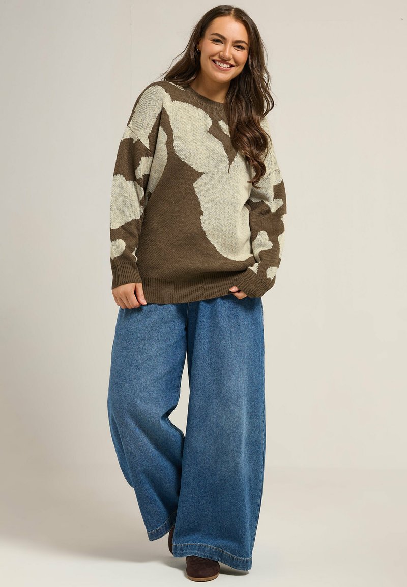 Przytulny, przerośnięty sweter w kolorze brązowym z kremowym, abstrakcyjnym wzorem, zestawiony z szerokimi, niebieskimi dżinsami. Materiał wydaje się miękki i ma luźny krój.