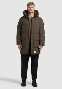 Zateplená parka v tmavě olivově zelené barvě s kapucí. Obsahuje zipové kapsy a nášivku s logem. Nosená přes hnědou košili a černé kalhoty. Černé boty.